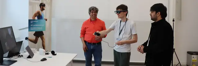 Three man standing at a table. One of them is wearing VR goggles