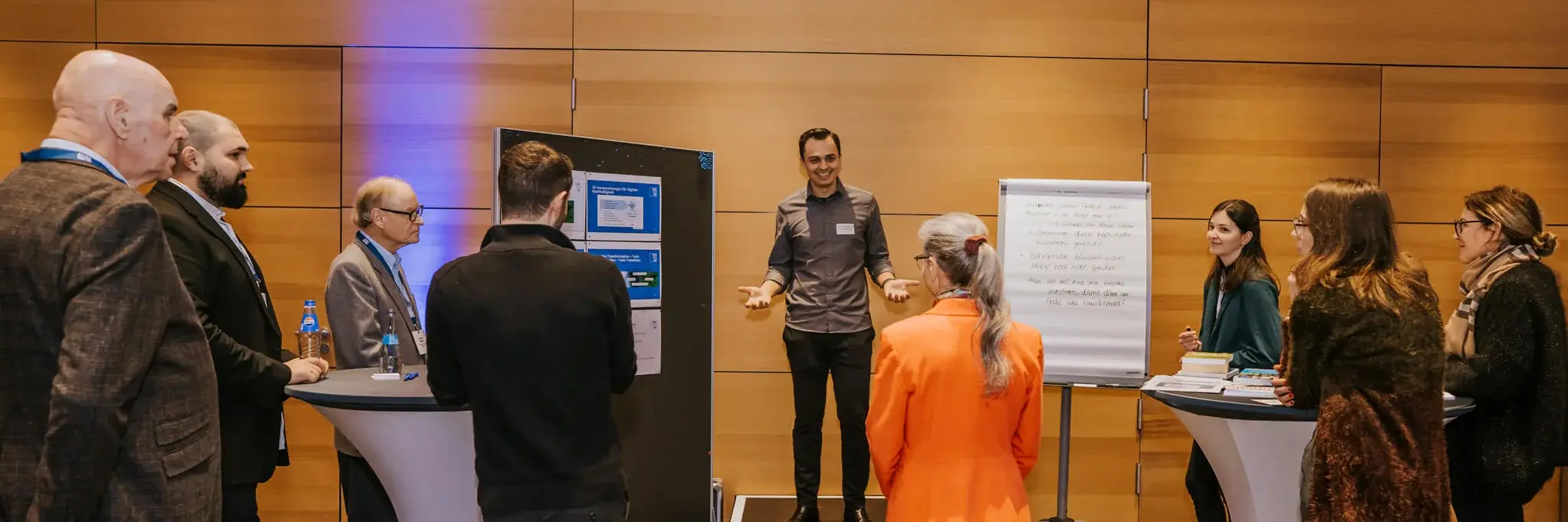 A group of people gathered around a speaker in a conference room. The speaker stands near a presentation board and flip chart. Attendees are dressed in business casual attire, listening attentively. The setting is modern with wooden panel walls.