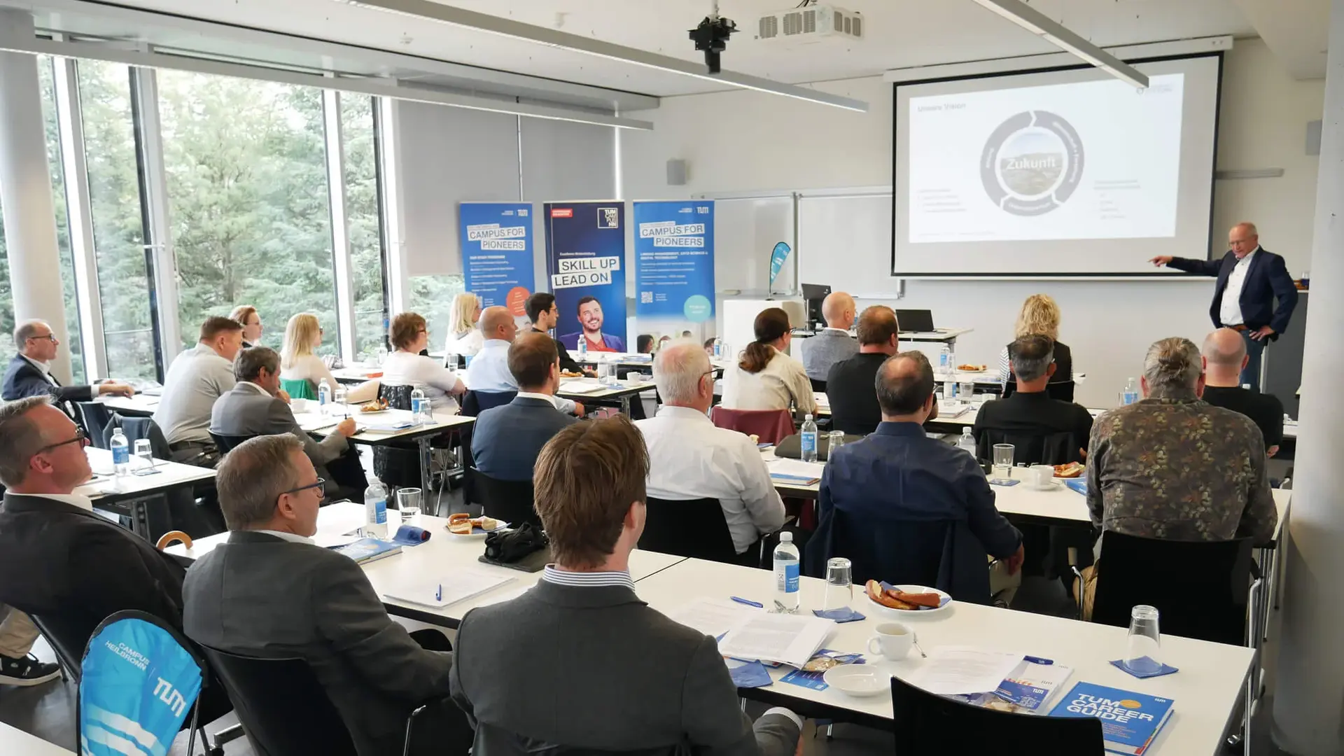 Eine Gruppe von Menschen sitzt in einem hellen Konferenzraum und hört aufmerksam einem Vortragenden zu, der auf einen Bildschirm mit einem Diagramm zeigt. Die Tische sind wie in einem Seminarraum angeordnet, mit Papieren, Wasserflaschen und Snacks.