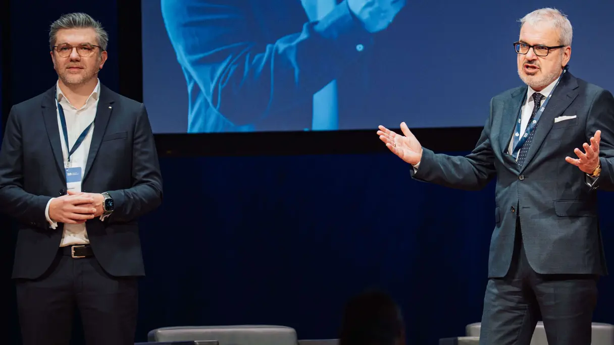 Three people stand on a stage in front of armchairs and a large screen displaying text. Two men are in suits; one woman in a purple suit is holding papers.