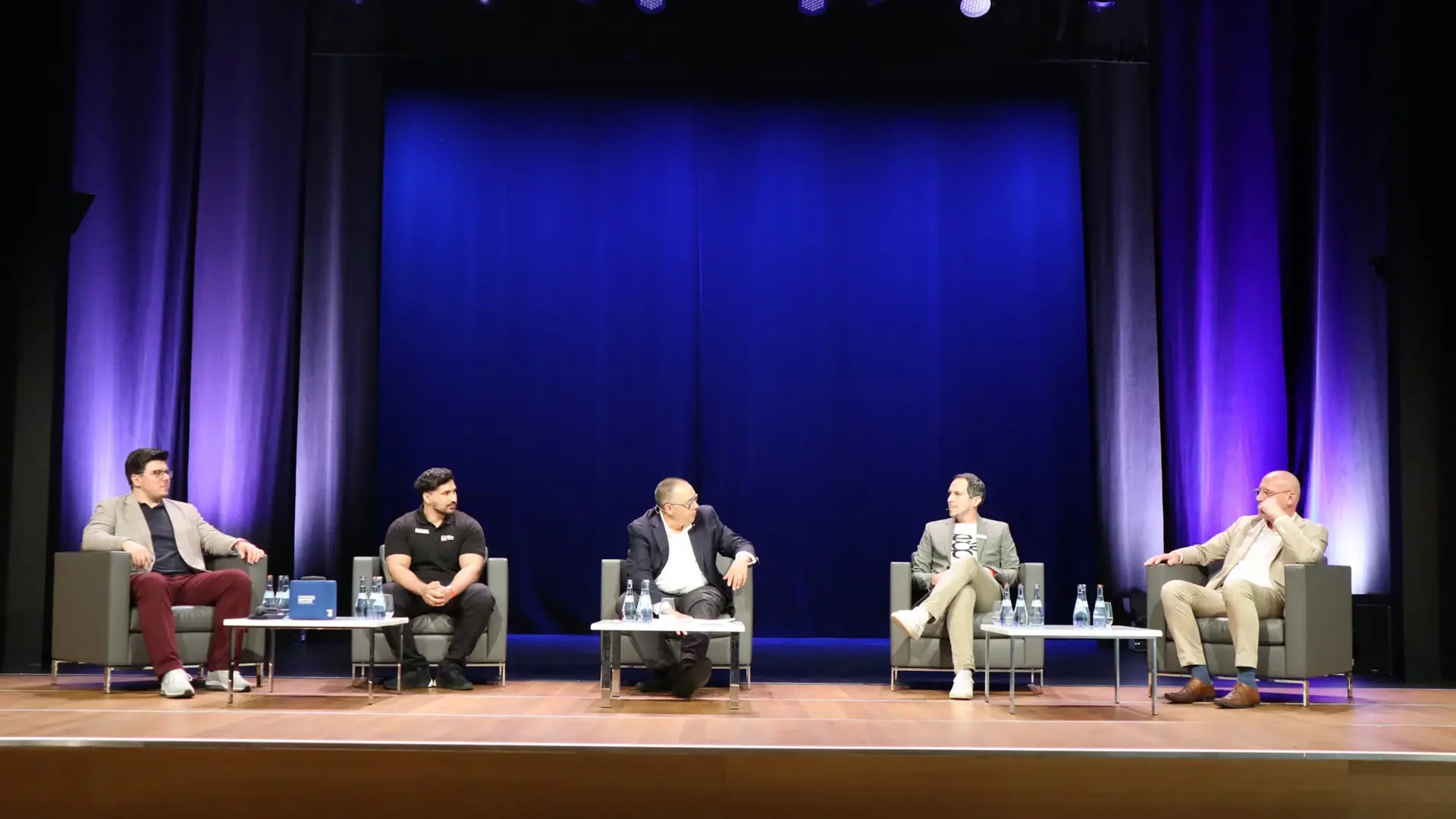 Five people are seated on a stage, engaged in a panel discussion. They are sitting in chairs, with small tables in front of them holding water bottles. The background features a blue lit curtain.