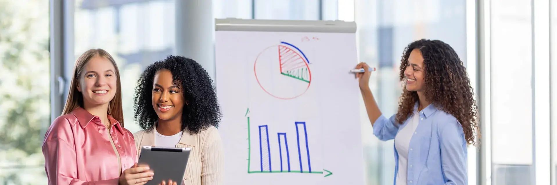 Drei Frauen befinden sich in einem Büro. Zwei von ihnen halten lächelnd ein Tablet in der Hand, während die dritte, in einem blauen Hemd, auf Diagramme auf einem Flipchart deutet. Die Diagramme zeigen ein Kuchendiagramm und ein Balkendiagramm. Im Hintergrund sind große Fenster zu sehen.