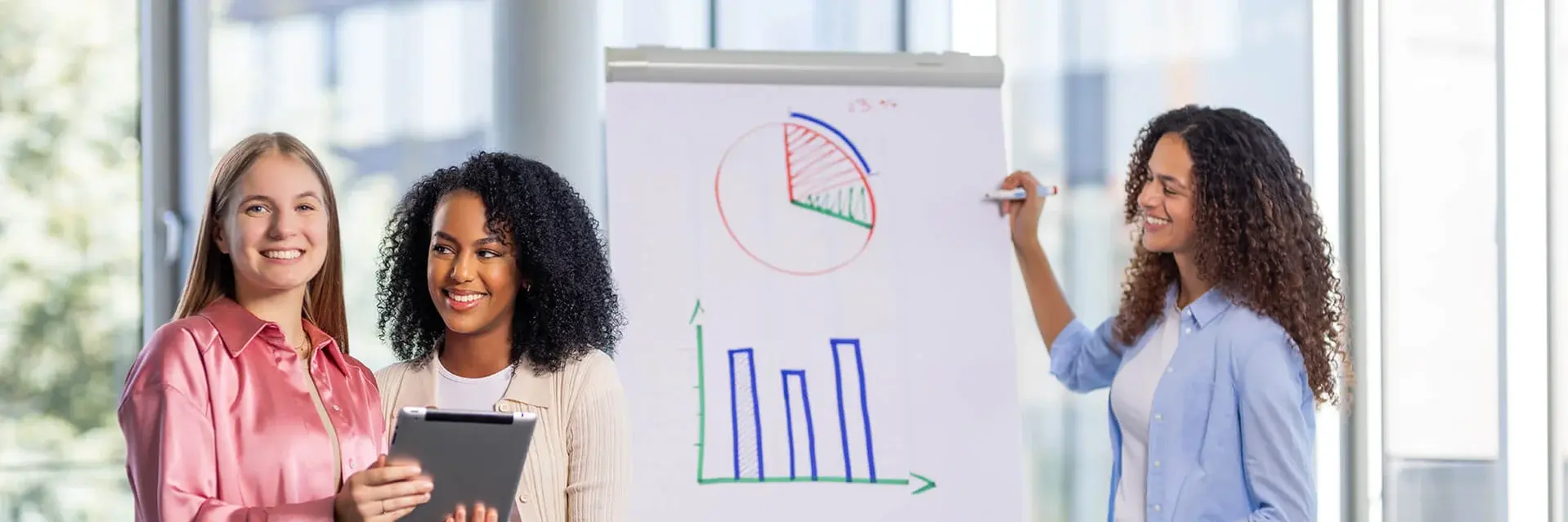 Three women are in an office setting. Two stand holding a tablet, smiling, while the third, in a blue shirt, gestures towards charts on a flip chart. The charts depict a pie chart and bar graph. Large windows are visible in the background.