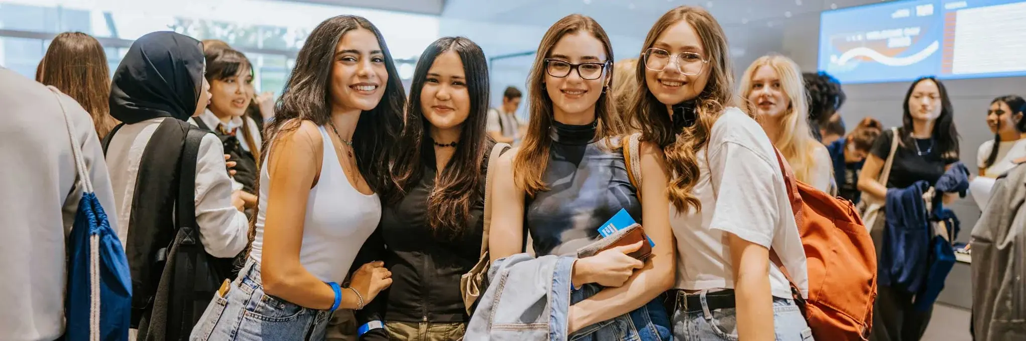 Eine Gruppe junger Frauen steht lächelnd in einer hellen, modernen Eingangshalle; sie posieren gemeinsam, tragen lässige Kleidung und halten Taschen oder Jacken.