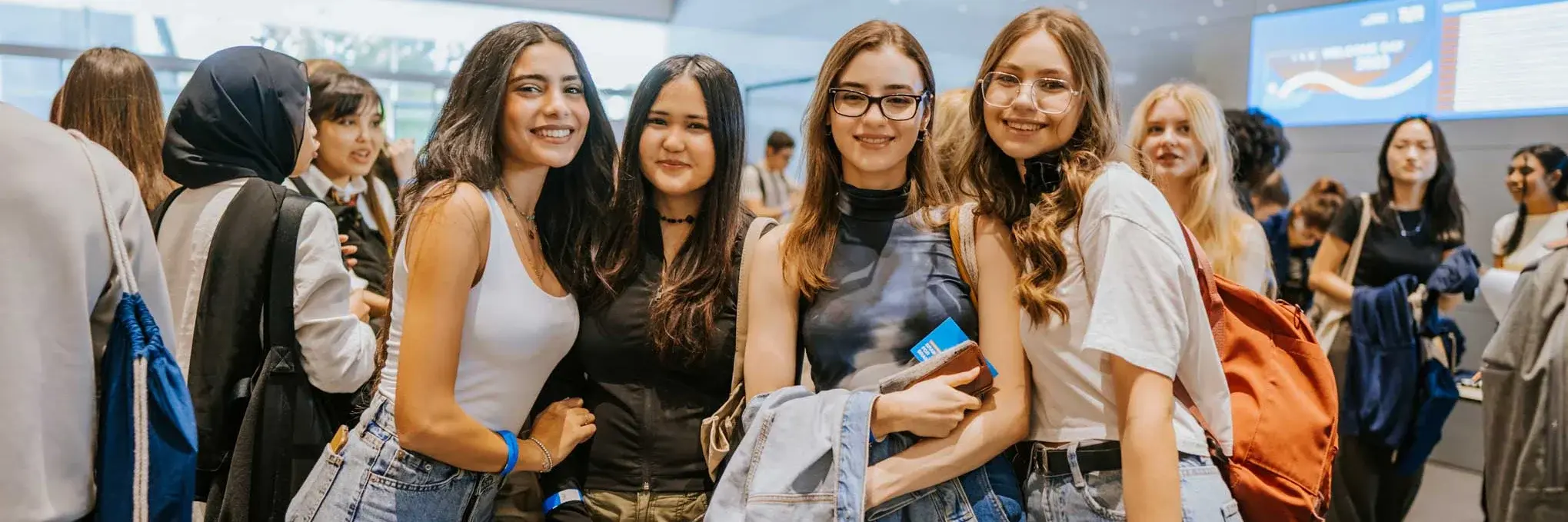 A group of young women smiling and posing together in a bright, modern lobby, dressed casually and holding bags or jackets.
