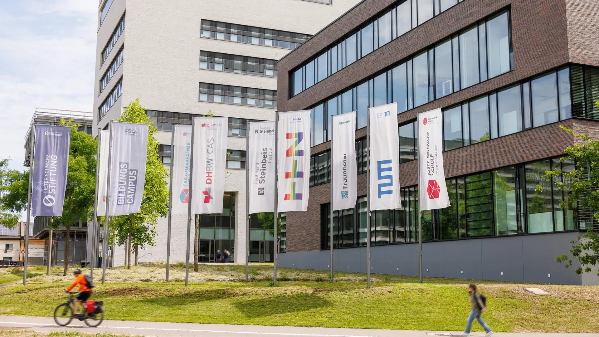 Flags of institutions at Bildungscampus Heilbronn in front of modern buildings, including TUM Campus Heilbronn, DHBW, Hochschule Heilbronn, Fraunhofer, Steinbeis, and others.