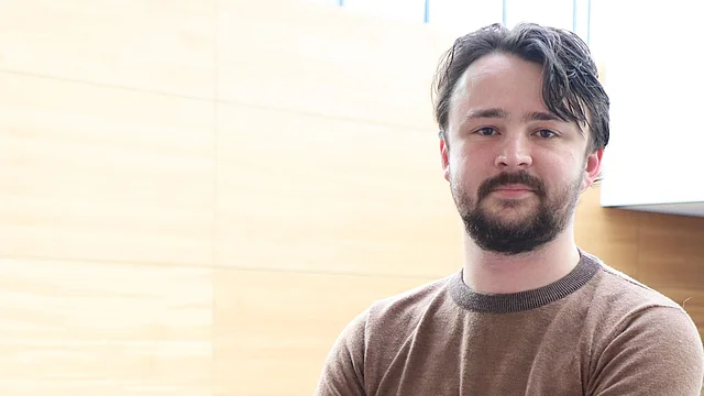 A man stands with crossed arms leaning on a railing in a modern interior with light wood paneling.