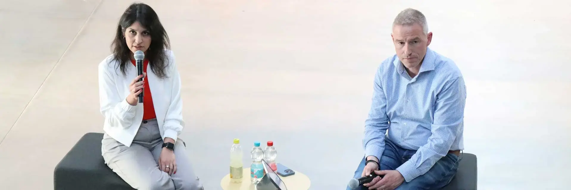 [Translate to German:] Two individuals seated on a stage, each with a microphone, engaged in conversation or discussion.
