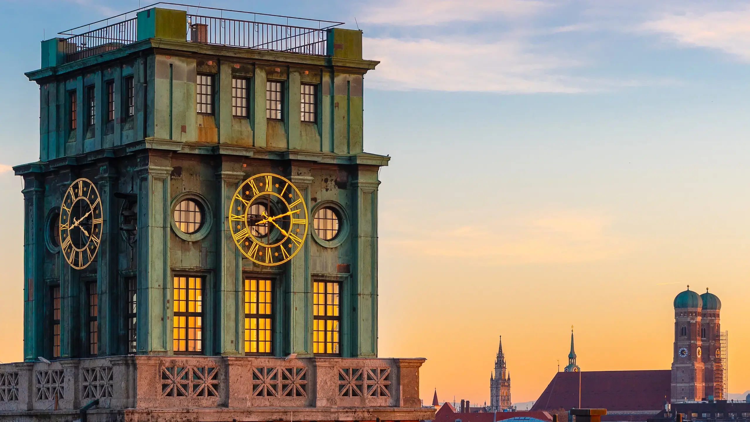 Ein grüner Uhrturm überragt bei Sonnenuntergang die Dächer, während in der Ferne der blaue Himmel und die beiden Kuppeln einer weiteren Kirche zu sehen sind.