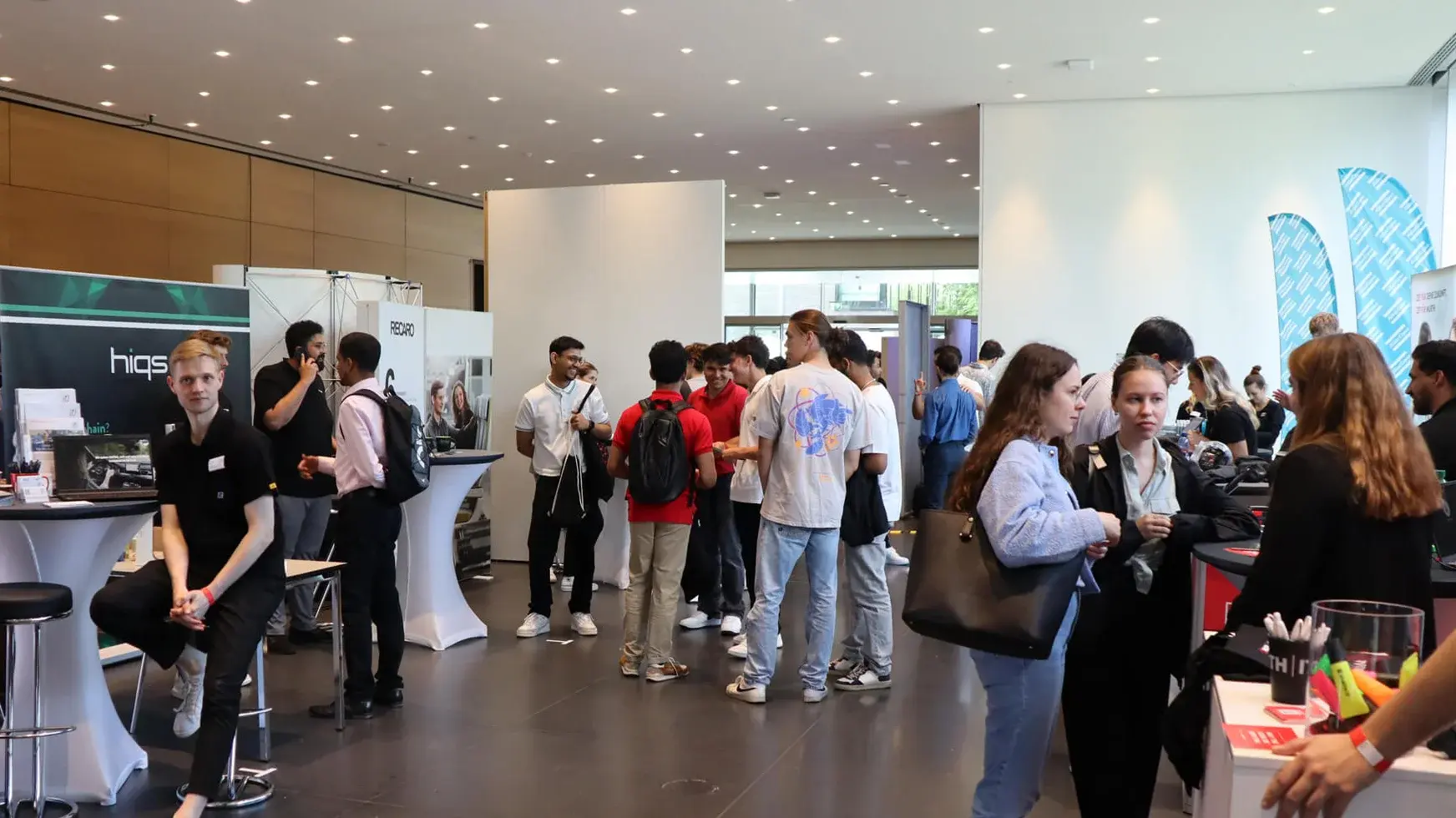 A modern, well-lit conference hall filled with people interacting at booths. Large screens and banners display company logos. Attendees are engaged in conversations, with some seated at high tables and others standing.