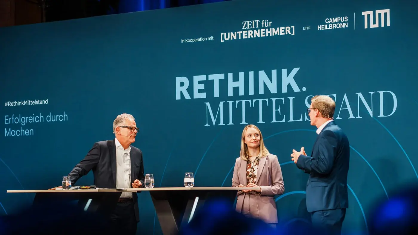 Three people stand at a high table on stage at the Rethink.Mittelstand event by TUM Campus Heilbronn, with the text 'RETHINK. MITTELSTAND' in the background.