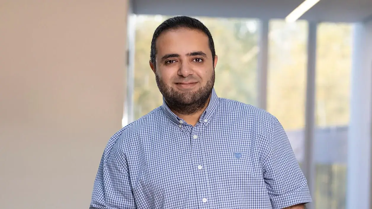 Prof. Amr Alanwar lehnt sich mit seinem linken Arm an ein Geländer in einem hellen Flur und trägt ein kurzärmeliges blau kariertes Hemd und eine blaue Hose.