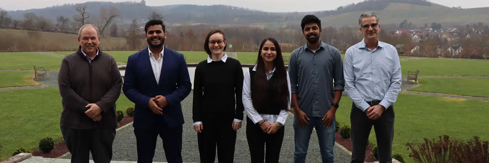 A group of six people standing outdoors on a patio with a scenic landscape in the background. They are dressed in formal and semi-formal attire.