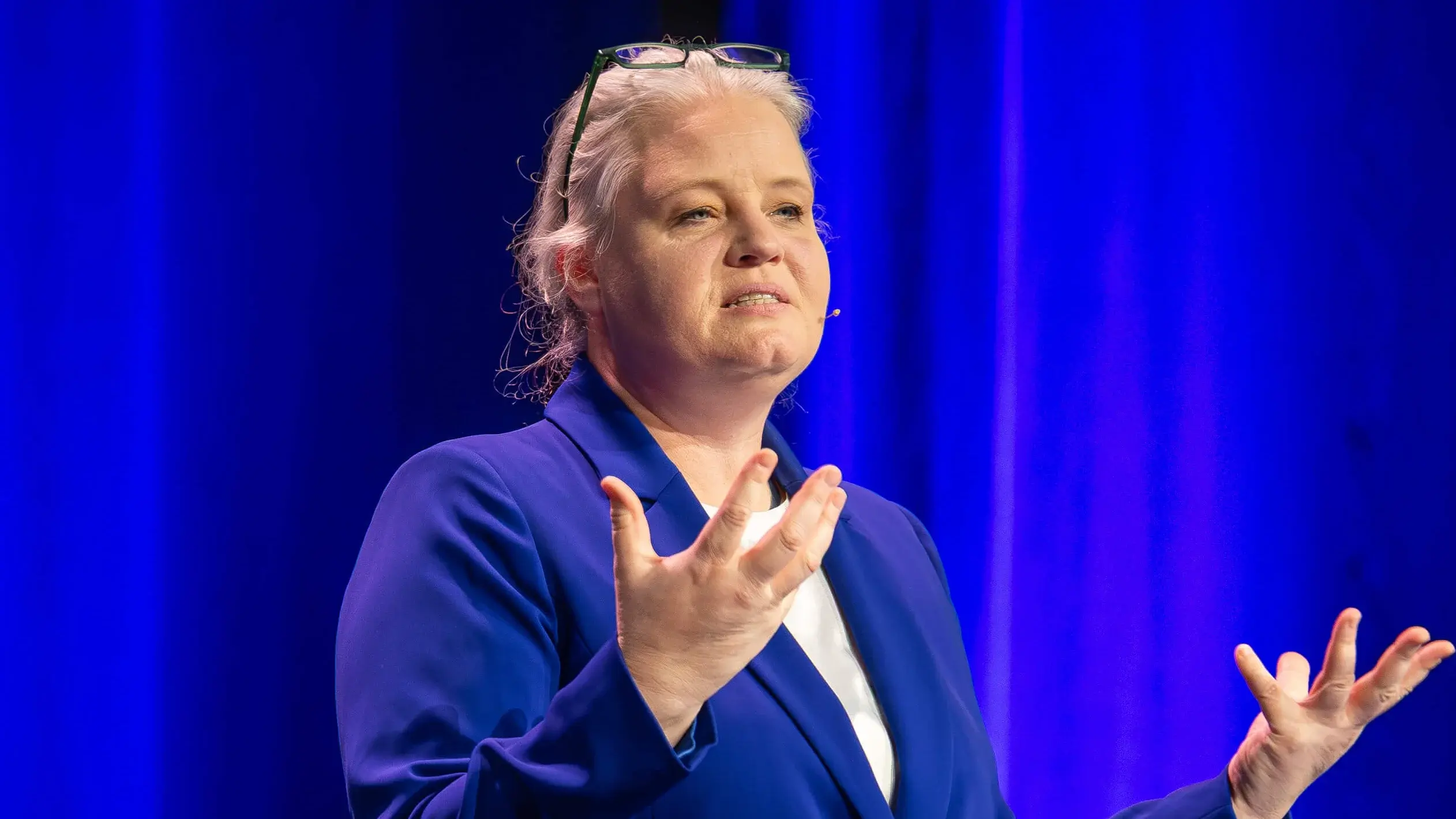 A person in a blue suit gestures while speaking on stage in front of blue curtains, standing next to a small round table.