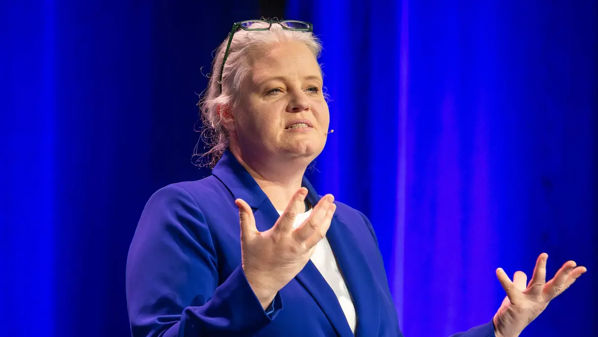 Eine Person in einem blauen Anzug gestikuliert, während sie auf einer Bühne vor blauen Vorhängen spricht und neben einem kleinen runden Tisch steht.
