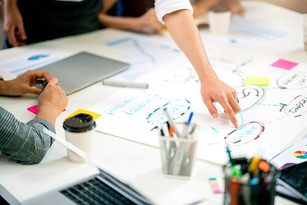 Several people collaborate around a table with charts, markers, a laptop, and coffee cups, discussing a project plan laid out on large paper.