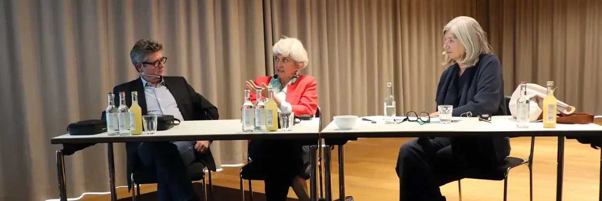 a woman und two men sitting behind a table, talking to each other