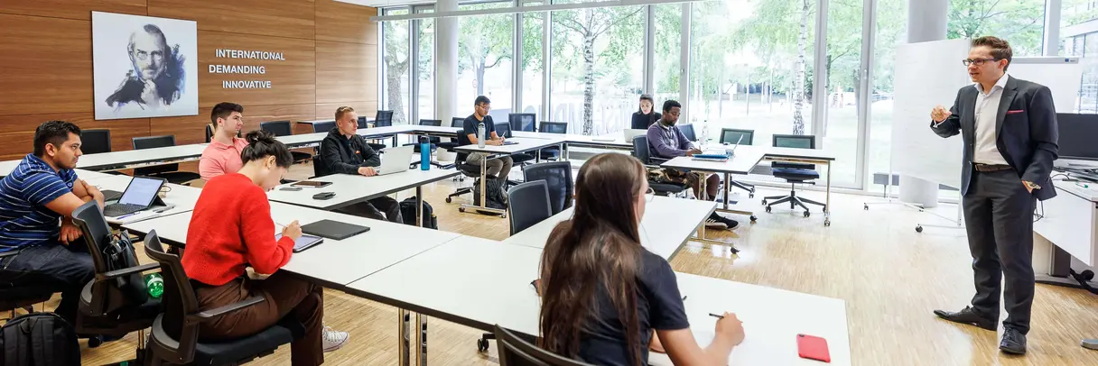 Eight students sit in a modern and inviting seminar room and listen attentively to a young lecturer dressed in a suit.