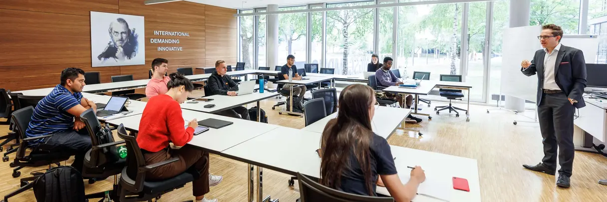 Acht Studierende sitzen in einem modernen und einladenden Seminarraum und hören interessiert einem jungen, in einen Anzug gekleideten Dozenten zu. 