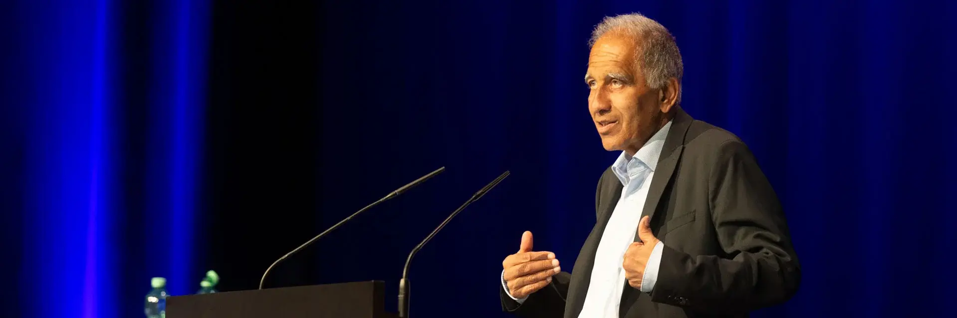 A man stands at a podium, delivering a speech to an audience, with a focused expression and gesturing with his hands.