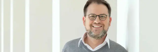 [Translate to German:] Man in gray jumper leaning against a white pillar
