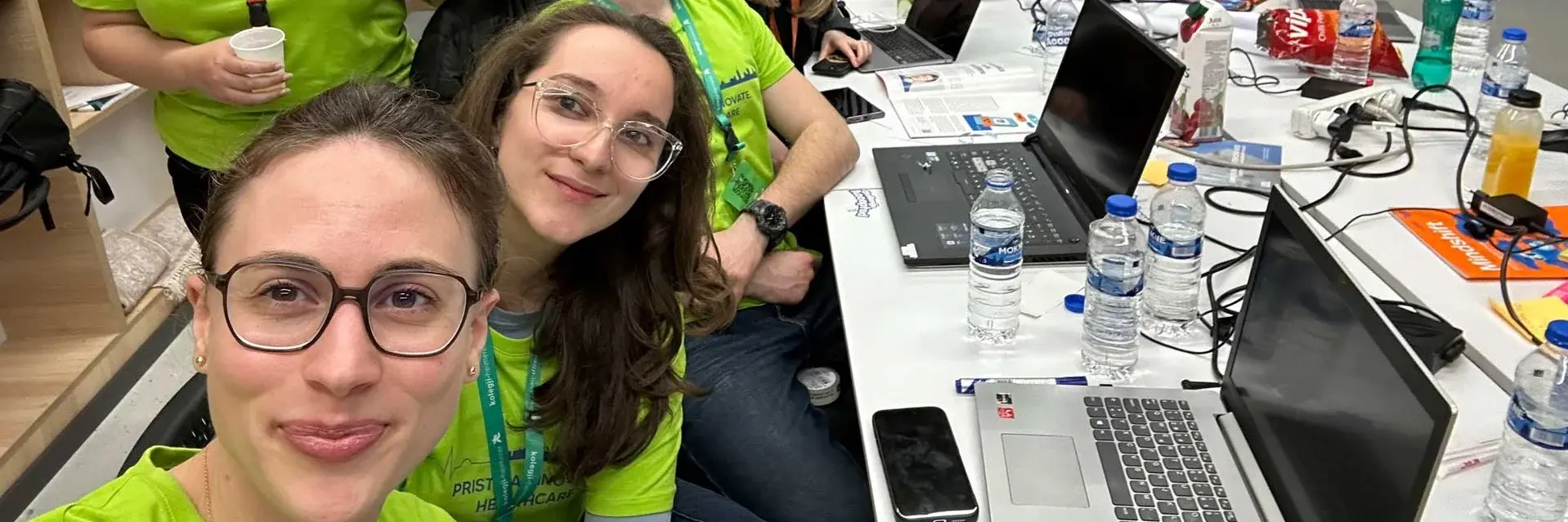 A group of eight people, wearing matching green shirts, pose around a table cluttered with laptops, water bottles, and snacks. They appear cheerful and are inside a room with a Sentry Cyber Solutions logo on the wall.