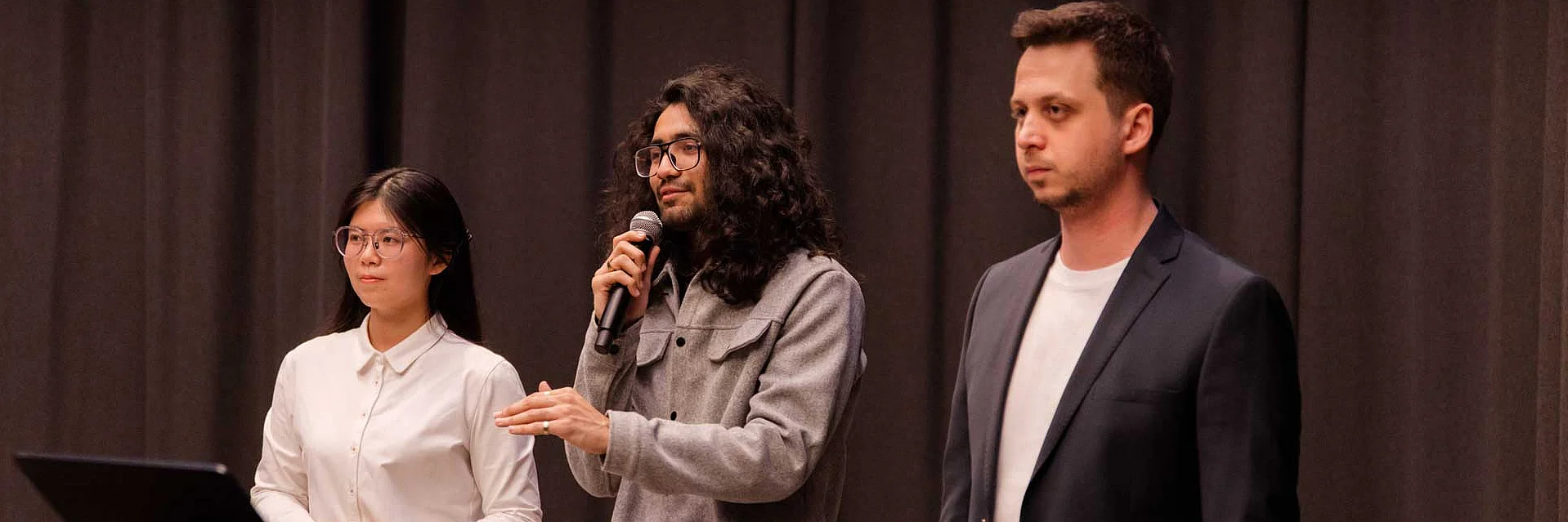 a woman and two men standing in row, the middle man speaking into a microphone
