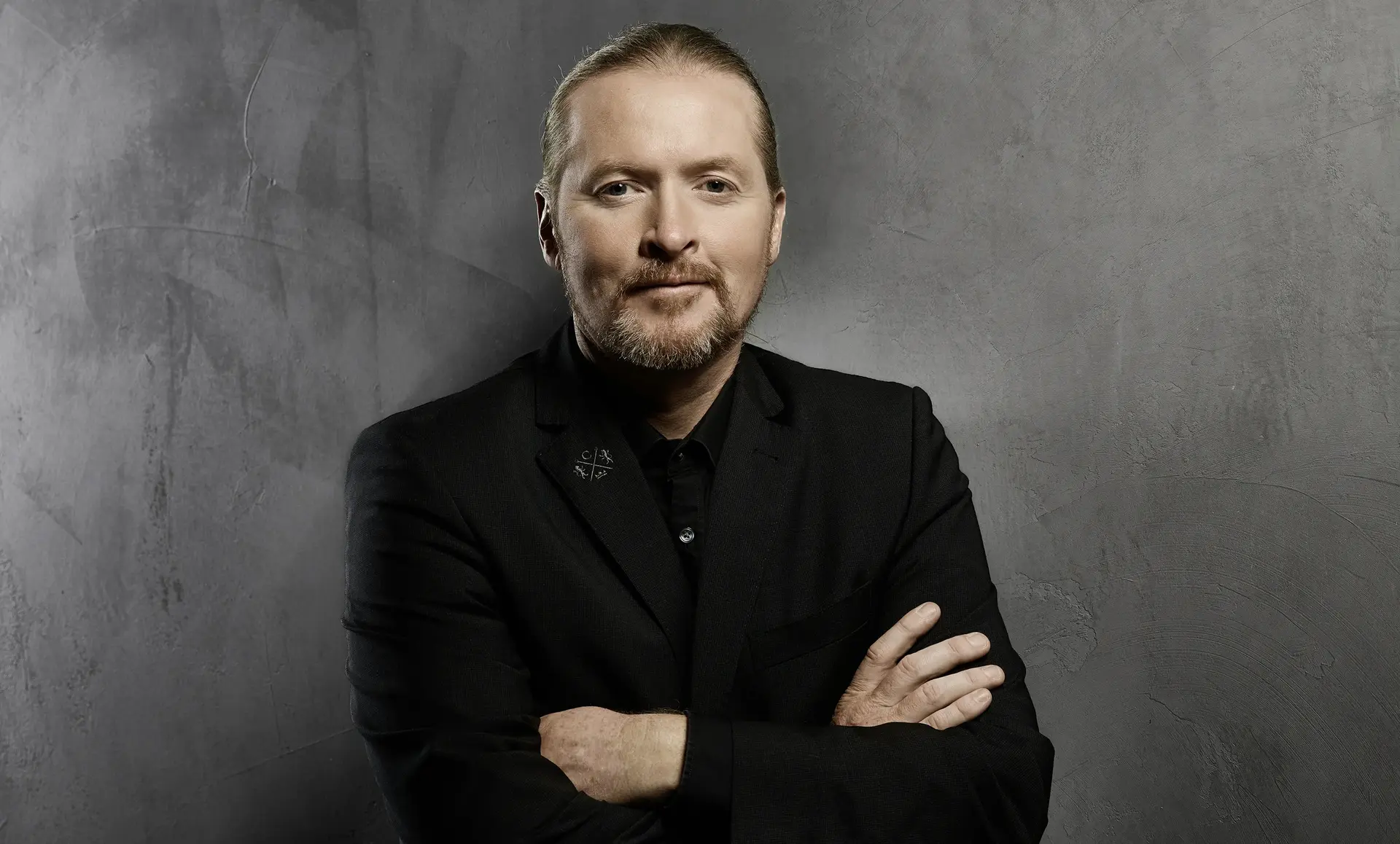 Joey Kelly in a dark suit stands with arms crossed against a gray wall.